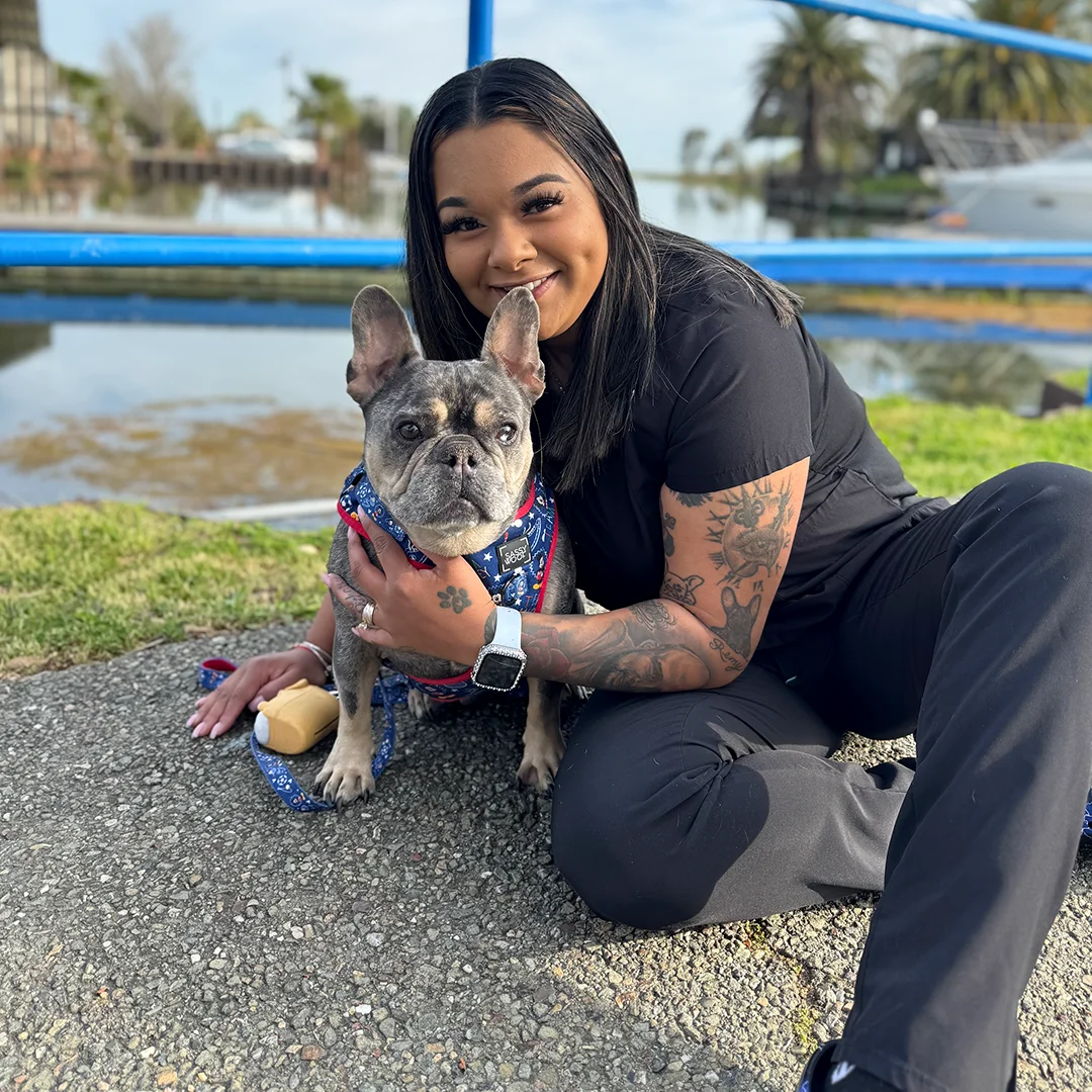 Smiling woman sitting on the ground hugging a gray French Bulldog in a patriotic bandana near a waterfront.