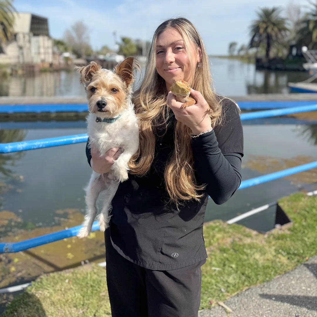 Smiling woman holding a small terrier mix in one arm and a bearded dragon lizard in the other near a waterfront.